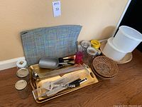 Wide view of lot contents showing placemats, trays, serving utensils, baskets, jars, and containers arranged against a wall on floor.