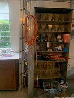 Photo showing two aluminum ladders (6-foot A-frame and 16-foot expandable) with orange extension cord hanging on ladder, metal cubby shelves filled with supplies, and a plastic bin on the floor.