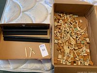 Box containing uncounted Mahjong tiles and domino sticks inside with three black wooden tile holders beside them.