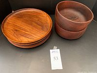 Five round wooden dinner plates stacked, natural wood grain visible in warm brown colors. Two dark wooden bowls stacked beside plates.