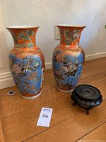 Photo of two tall ceramic vases and a wooden pedestal on hardwood floor against white wall, showing colors and design of the vases.