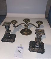 Front view of the pair of ornate Old English silver candlesticks and the three-arm silver candelabra on white backdrop.