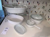Classic white CorningWare bakeware set displayed together: three ribbed exterior dishes with glass lids, an oval dish, a small handled pot with lid, and metal riser.