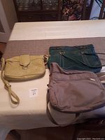 Three handbags displayed on a table: tan leather handbag, green leather handbag, taupe fabric travel bag