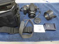 Panasonic Lumix DMC-FZ28 camera body with attached adjustable zoom lens, lens cap, lens hood, charger, and black carrying case displayed on blue cloth background.