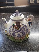 Front side view showing the white ceramic teapot decorated with purple floral patterns, daisy flower on base, and cut-out windows with animated figures inside.