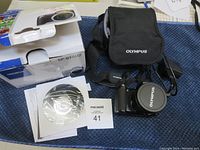 Olympus SP-810UZ camera with lens cap, soft carrying case, original box, manual and DVD disc displayed on a table.