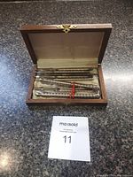 Open wooden box displaying a metal nutcracker and several metal nut picks arranged inside on a beige fabric lining under lid with a brass clasp on the front of the box.