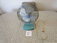 Photo of vintage table fan showing fan from a diagonal front angle with power cord coiled beside base on a patterned surface