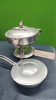 Wide view of the hammered aluminum chafing dish with stand and lid along with small hammered pan with handle and lid on display.