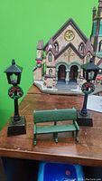 Photo of two black battery-operated lamp posts, a green metal park bench, and a hand painted porcelain church with gothic architectural details.