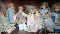 Group photo of eight porcelain collectible dolls in various outfits and sizes, arranged close together on shelf, featuring both early 1900s and more modern dolls.