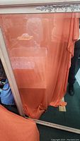Full view of the large wall mirror with an orange fabric placed behind it to reduce reflections. Shows beveled glass and whitewash wood frame with floral decoration.