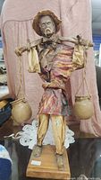 Full view of handcrafted paper mache man figurine with ceramic jugs, showing mounted wooden base and detailed clothing and facial features.