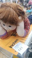 Close-up front view of Musical Lin Doll face and upper body, showing red hair in pigtails with blue hairbands wearing a red patterned top, sitting on a yellow measuring board.