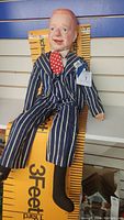 Full view of WC Fields dummy doll sitting against a wall, wearing blue pinstriped suit, red polka dot tie, no shoes, one sock torn.
