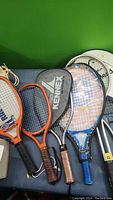 Six racquets laid out, showing three Pro Kennex racquetball racquets with distinct orange and black frames and leather grips, with visible strings and branding.