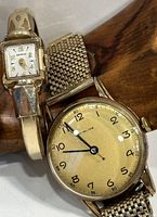 Pair of two vintage Benrus watches displayed side-by-side on wooden surface. Ladies watch features rectangular face with white dial and gold numerals, men's watch has round face with yellowish dial and black numerals. Both show metal bands.