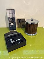 Photo showing the boxed wall-mount wine opener and stopper set, wooden ice bucket with lid, and boxed lever-style wine opener set on a green surface.