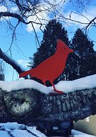 Red powder-coated steel cardinal standing on snow-covered tree branch outdoors in winter, showing full silhouette against sky and trees.