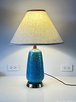 Full view of the lamp lit on a white surface showing ceramic body, teak neck, metal base, and linen shade.