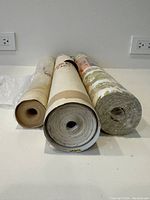 Overall view of the three wallpaper rolls placed horizontally, showing the ends and paper edges, appearing new with one partially used.
