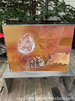 Full view of framed abstract mixed media artwork showing warm earth tones and textures on an outdoor background