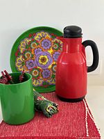 Photo showing the vintage POP flower metal tray with colorful floral pattern, red Swedish thermos, green utensil holder with red-tipped utensils, and cotton placemats arranged on textured red mat.