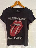 Front view of black T-shirt hanging on wooden hanger, showing The Rolling Stones lips and tongue logo in red, text above and below the logo reading 'THE ROLLING STONES 1975 - US TOUR'.