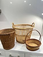 Three vintage wicker baskets of different sizes displayed on a white surface with a neutral background. Includes one large open-weave basket with handles, one medium tall woven basket, and one small round basket with a handle.