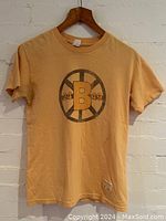 Full front view of T-shirt hung on wooden hanger, showing orange-beige color and Boston Bruins logo with 24 and 49 on wheel design.