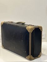 Full view of the vintage black suitcase showing rectangular shape, handle, and worn protective corner caps