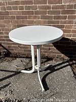 Front view of the round white metal garden table showing the solid metal top and tubular legs against a brick background.