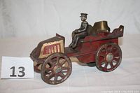 Side angle view of the antique toy car showing red and gold paint, four spoked wheels, and the seated driver figure.