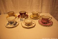 Six vintage teacups and saucers displayed in a row on table, showcasing variety of pattern and colors, including gold, pastel yellow and pink, white with blue floral, and others