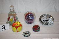 Six glass items on white cloth background: bell-shaped paperweight, round yellow flower paperweight, red-white-blue swirly paperweight, black-white patterned paperweight, miniature signed paperweight, and round art glass drawer pull.