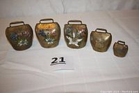 Five vintage Swiss steel souvenir bells with brass finish and hand-painted flowers arranged on a white cloth showing their relative sizes.