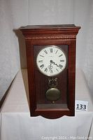 Full view of rectangular wooden wall clock with glass door, pendulum visible, placed on white cloth.