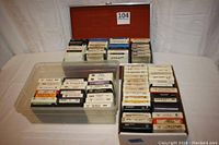 Overall view of three containers holding numerous 8-track tapes, with a wood carrying case at rear and plastic bin with 24 tapes in front left, and cardboard box with tapes front right.
