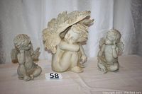 Three angel figurines of varying sizes and postures displayed on a white surface against a curtain background.