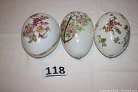 Three vintage Easter eggs shown together on white cloth background. The eggs have floral and Easter-themed hand-painted decorations, with one porcelain marked West Germany two-piece egg.