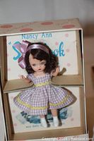 Storybook doll with purple gingham dress, white shoes, purple ribbon, eyes closed, in box.