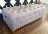 Side angled view showing tufted button design, silver-tone handle on the side, wooden block legs, and fabric texture.