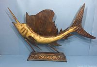 Full view of the metal swordfish sculpture showing its textured yellowish-gold body and brown fins, mounted on a metal base with wave design.