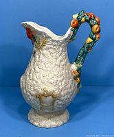 Side view showing the textured white body with painted wheat and floral decoration, and fruit-adorned handle.