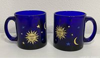 Two cobalt blue glass mugs side by side showing gold sun face surrounded by moons and stars
