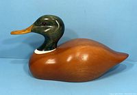 Side view of the carved wooden mallard decoy showing the brown body, green head, and orange beak against a blue background.
