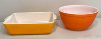 Two vintage Pyrex dishes side by side: an orange mixing bowl and yellow rectangular baking dish.