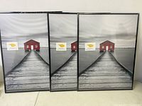 Three 18x24 black picture frames standing side by side, front view showing the art print of a dock leading to a red house.
