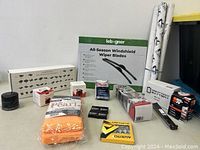 Overview of automotive items arranged on floor including windshield wiper blades, oil filters, car battery, spark plugs, microfiber towels, and wrapped decals.